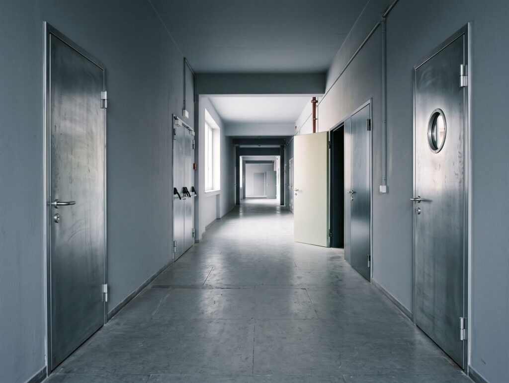 Office corridor, one person unlocking a door with a mechanical key, another walking down the corridor with a laptop, both wearing ID badges on lanyards.